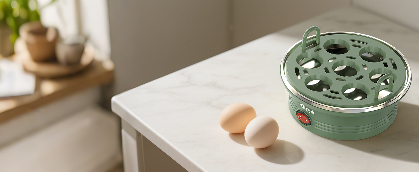 Green electric egg cooker with perforated lid on white countertop. Two eggs placed nearby. Kitchen setting with wooden accessories visible in background.