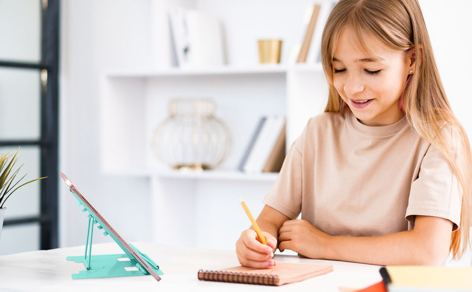 Young girl writing in notebook at desk with teal tablet stand. White shelving unit with decorative items in background.