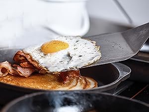 Frying pan on stovetop with sunny-side up egg and bacon being cooked. Metal spatula visible in frame.