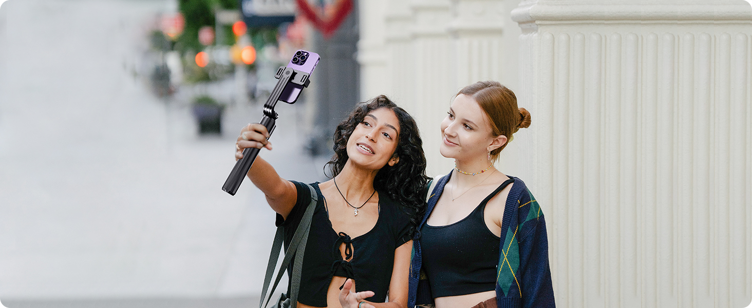 A handheld selfie stick with a smartphone attached, being held up by an arm. The device is extended outwards, demonstrating its use for taking group photos.