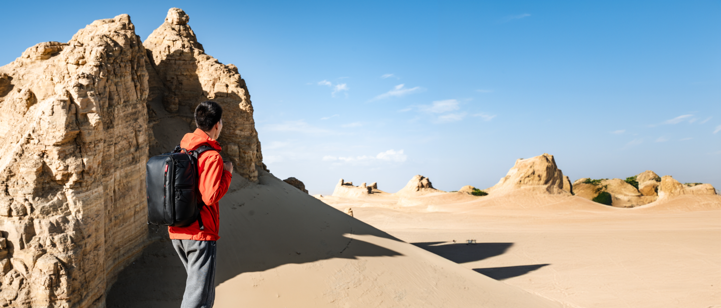 A hiker with a dark backpack stands beside a tall rock formation in a desert landscape with scattered rock formations against a blue sky.