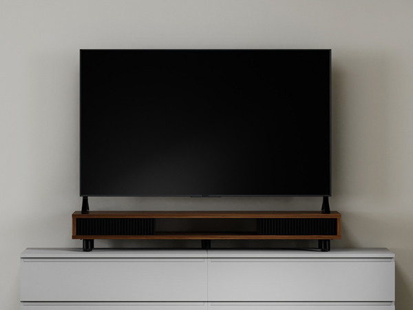 Large black rectangular electronic device, possibly a TV or sound bar, mounted on a wooden stand atop a white cabinet against a light-colored wall.