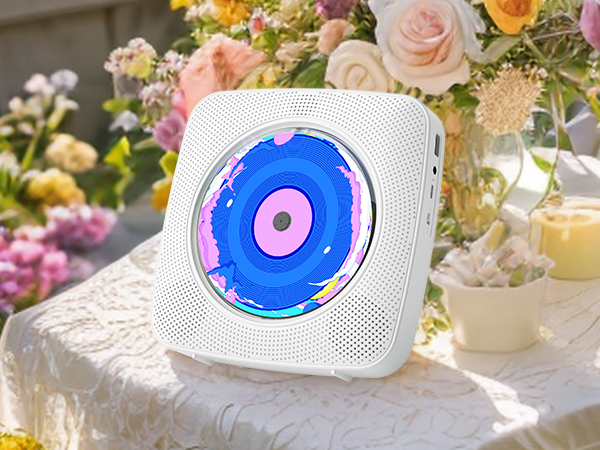 White square device with colorful circular display, featuring blue and pink concentric circles. Sits on lace tablecloth near floral arrangement and candle.