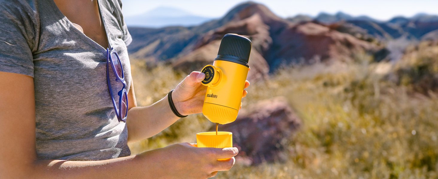 Hand holding a yellow handheld device against a desert landscape background. The device appears to be a compact outdoor tool or instrument.