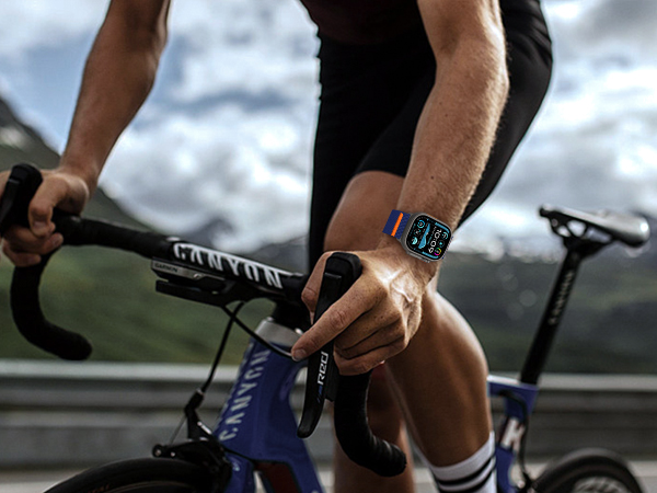 Cyclist gripping handlebars of blue racing bike, wearing smartwatch with digital display. Mountain scenery visible in background.