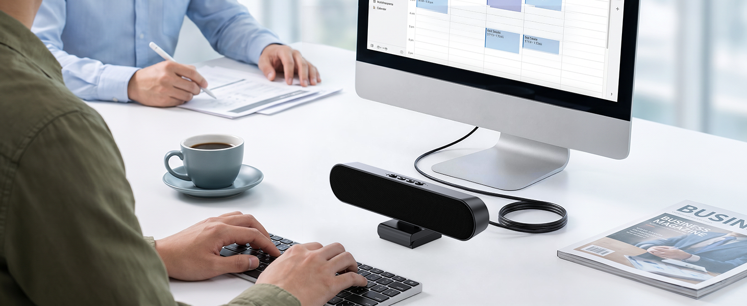 Office workspace scene showing person's hands at desk with laptop, tablet, pen holder, coffee cup, and wireless speaker on white desk surface with window background.