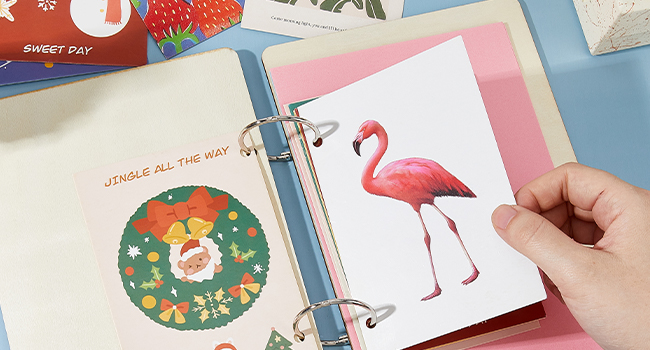 Open ring binder with colorful pages. One page shows a Christmas wreath illustration, another displays a pink flamingo drawing. A hand is visible turning a page.