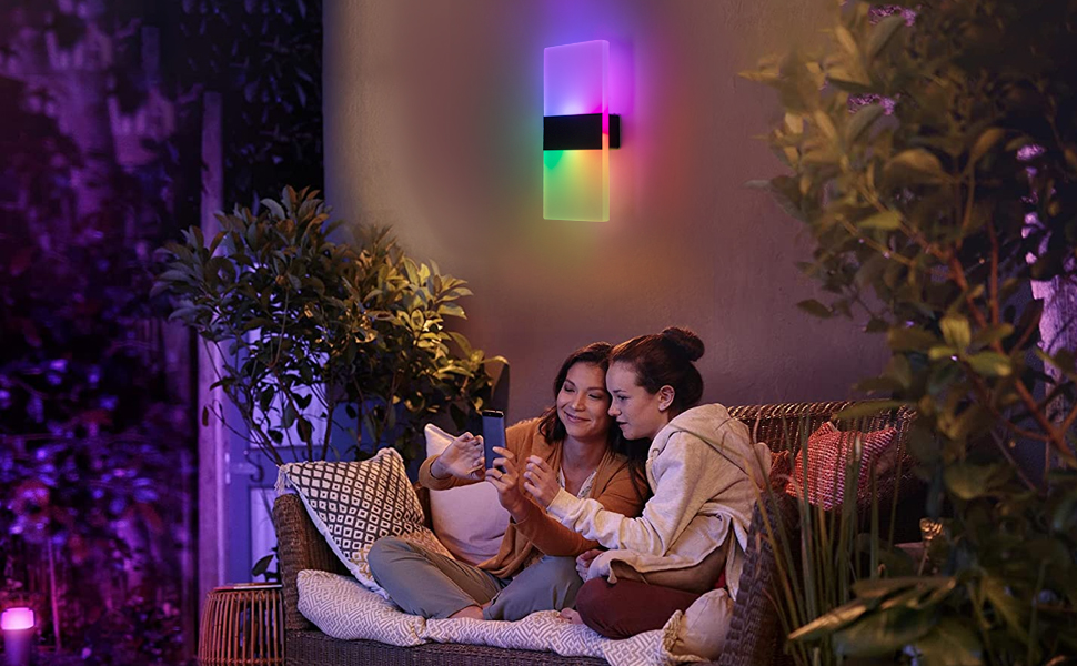 Colorful smart light fixture on wall, illuminating outdoor seating area with plants. People sitting on couch using electronic device.