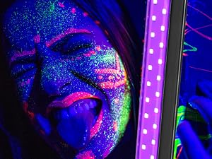 Close-up of face covered in fluorescent body paint. Vibrant blue, pink, and purple colors under UV lighting. Glitter effect visible on skin.