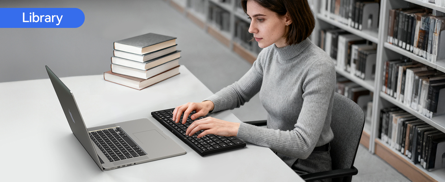 Text reads "Library". Person in gray shirt working at laptop beside stack of books on white desk in library with bookshelves visible in background.