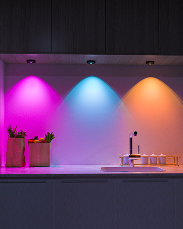Kitchen under-cabinet lighting system with three color options: pink, blue, and orange. Lights illuminate countertop with potted plants and kitchen items.