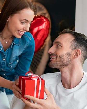 Couple exchanging red gift box, woman in blue denim shirt and man in white shirt with red heart-shaped balloons in background.