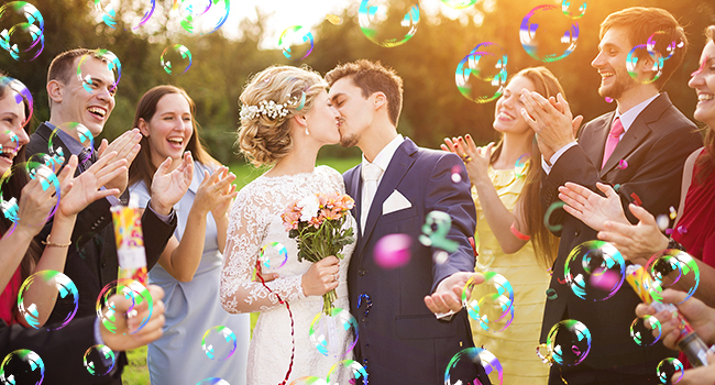 Outdoor wedding scene with newlyweds kissing, surrounded by guests. Colorful soap bubbles fill the air, creating a festive atmosphere.