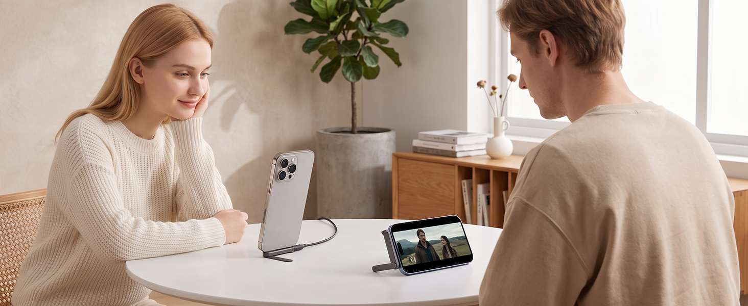 Two people at a table in a bright interior space with potted plant. Person on left holds smartphone while person on right gestures during conversation, demonstrating device use.