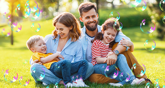 Family sitting on grass with soap bubbles floating around them. Two adults and two children smiling and embracing in a sunny outdoor setting.