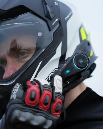 Motorcycle helmet-mounted communication device with blue light and control button. Rider wearing full-face helmet and red-accented gloves holds the device near their ear.