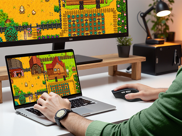 Desktop setup with monitor and laptop displaying colorful pixelated farm game. Desk has wooden stand, plant, and lamp. Person's hands on laptop keyboard, wearing watch.