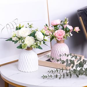 Two vases with floral arrangements on a white surface. One vase is white with white and pink flowers, the other is pink with white flowers.