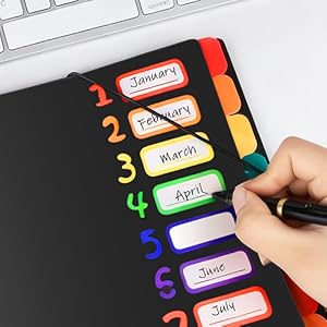Text reads 'January, February, March, April, June, July'. Rainbow-colored numbered tabs on black surface with handwriting being added to labels. Calendar or planner organization system.
