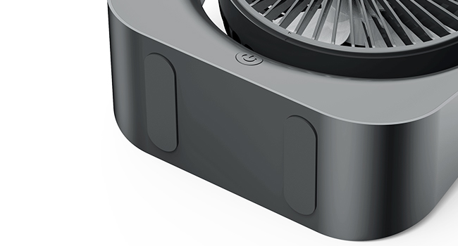 Close-up of a dark gray electronic device base with rounded edges. A circular fan or vent is visible on top, along with a small button or indicator light.