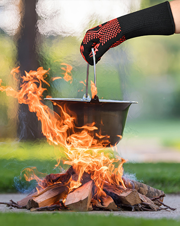Heat-resistant glove with red silicone grip pattern holding tongs over a flaming campfire. A black cooking pot is suspended above the fire.