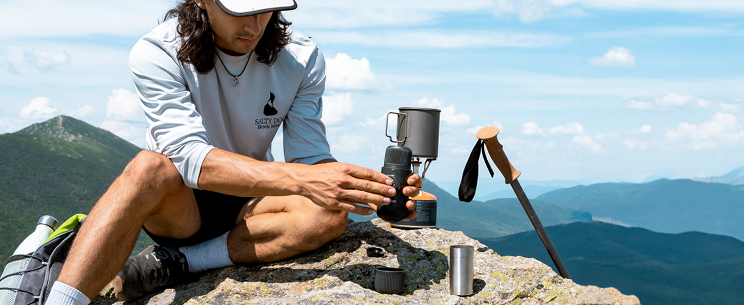 Person using portable coffee maker on rocky mountaintop. Scenic view of mountains in background. Demonstrates outdoor use of compact brewing device in nature setting.