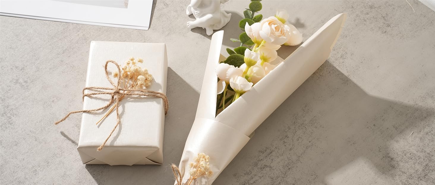 Gift wrapping accessories on concrete surface: white wrapped present with twine bow, cream paper cone holding flowers, and dried floral elements.