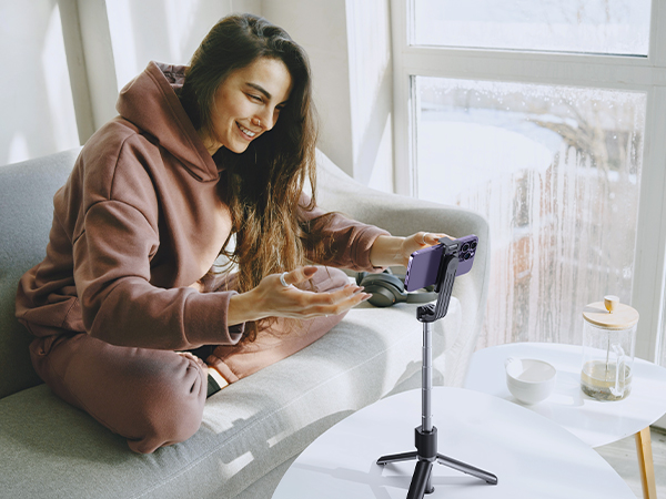 A smartphone tripod stand with adjustable height and angle, shown in use with a person recording or video chatting. The stand has a black frame and purple accents.