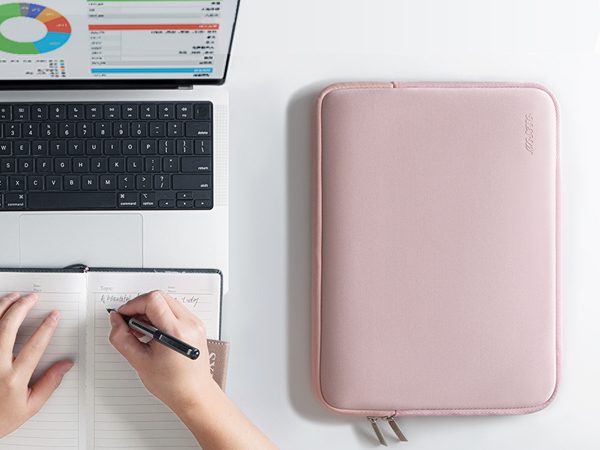 Pink rectangular zippered tablet or laptop sleeve next to a keyboard, notebook, and hands writing. Sleeve has a soft, textured surface.