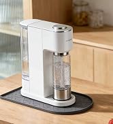 Compact white carbonated beverage maker with metallic silver dispenser head, positioned on black stand atop wooden countertop. Two filled glasses with ice and beverages visible beside the machine.