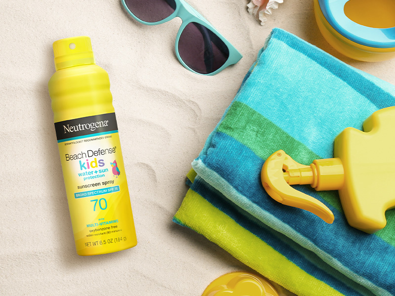 Neutrogena Beach Defense Kids Sunscreen Spray laying on the sand next to beach gear