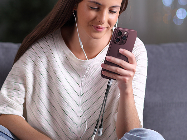 Woman wearing light gray cable-knit sweater holding burgundy smartphone, seated indoors with white earbuds around neck.