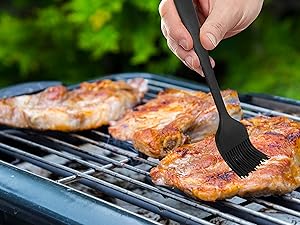 Close-up of hand brushing marinade on chicken pieces grilling on barbecue grate. Green foliage visible in background.