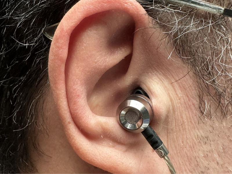 Close-up of ear wearing small metal wired earbud with mesh detail and silver cable