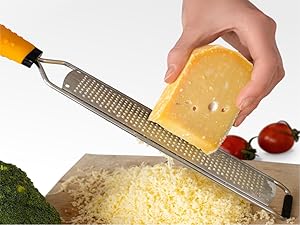 Stainless steel cheese grater with yellow handle being used to grate yellow cheese over a pile of grated cheese. Broccoli and tomatoes visible.