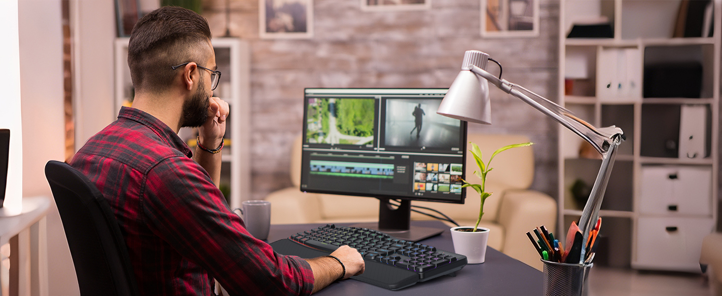 Person seated at desk wearing burgundy shirt, working at computer setup with dual monitors displaying video editing software in home office with shelving.