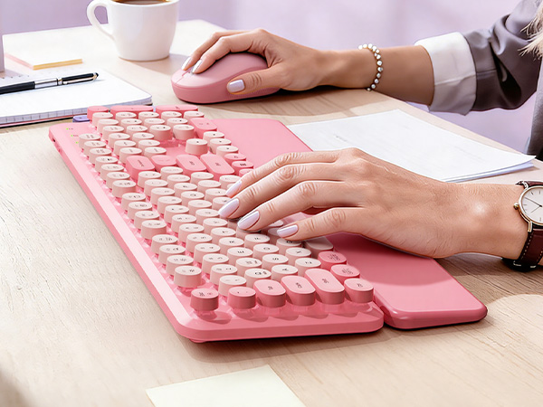 Woman typing on a Blush Pink retro wireless keyboard with matching mouse on a bright office desk.