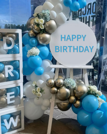 Birthday party decoration with blue and gold balloons, white flowers, and a circular sign reading 'HAPPY BIRTHDAY' in blue text. White cube shelves spell 'DREW'.