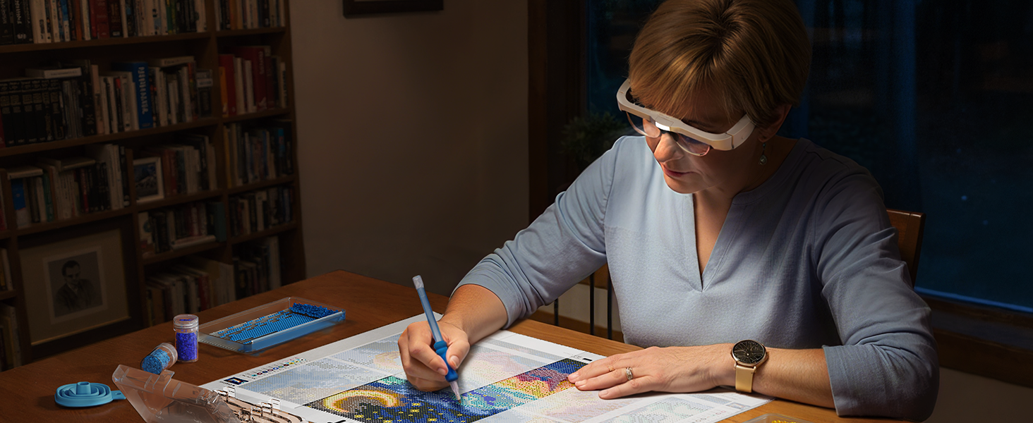 Person writing or drawing at a desk in dim lighting, surrounded by bookshelves in background.