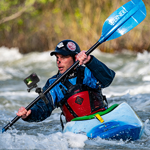 Action photograph of person in blue jacket and black cap paddling blue kayak on white water rapids, holding blue paddle, wearing safety gear.