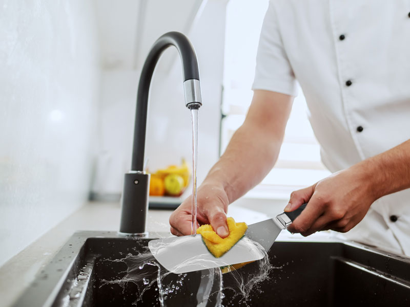 Black kitchen faucet with pull-down sprayer shown in use while washing a yellow dish in stainless steel sink.