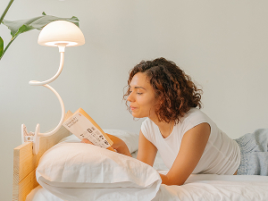Person relaxing in bed with white bedding while holding a round cookie or biscuit, with a table lamp visible in background.