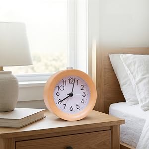 Wooden round alarm clock with blue and purple face displaying numbers displayed on bedside table with lamp, books, and white pillows in bedroom setting.