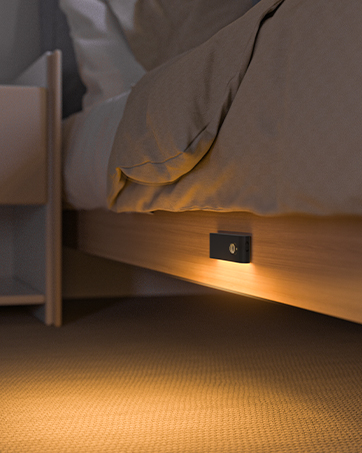 Small wall-mounted motion sensor night light casting a warm amber glow on carpet beneath a bed frame, providing subtle floor illumination.
