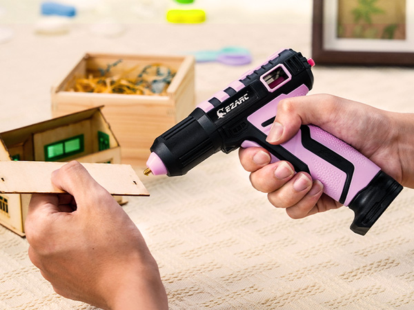 Black and pink hot glue gun being used for crafting, with wooden materials visible in background and a storage box containing supplies.