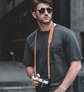 A person wearing sunglasses and a dark t-shirt holds a camera with an orange strap in an urban setting.