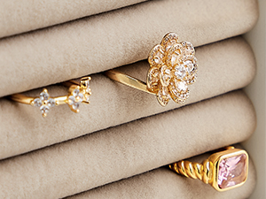 Four gold rings displayed on beige fabric. Designs include floral patterns, gemstones, and textured bands.