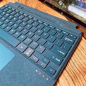 Close-up of a blue laptop keyboard with fabric-covered palm rest. Keys are backlit, and a partial view of the trackpad is visible. The device appears to be on a wooden surface.