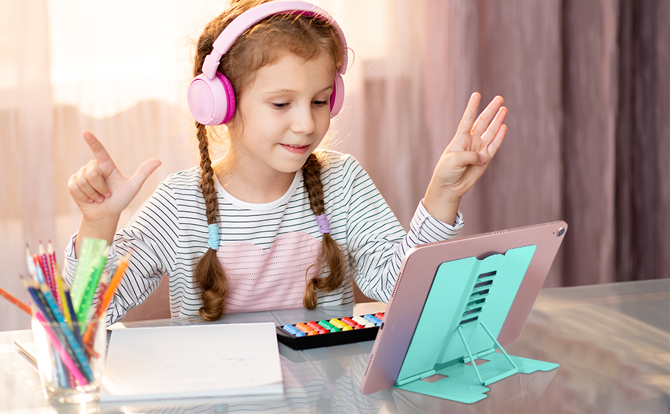 Child wearing pink headphones using a tablet in mint green stand. Colorful pencils in cup nearby. Striped shirt, braided hair.