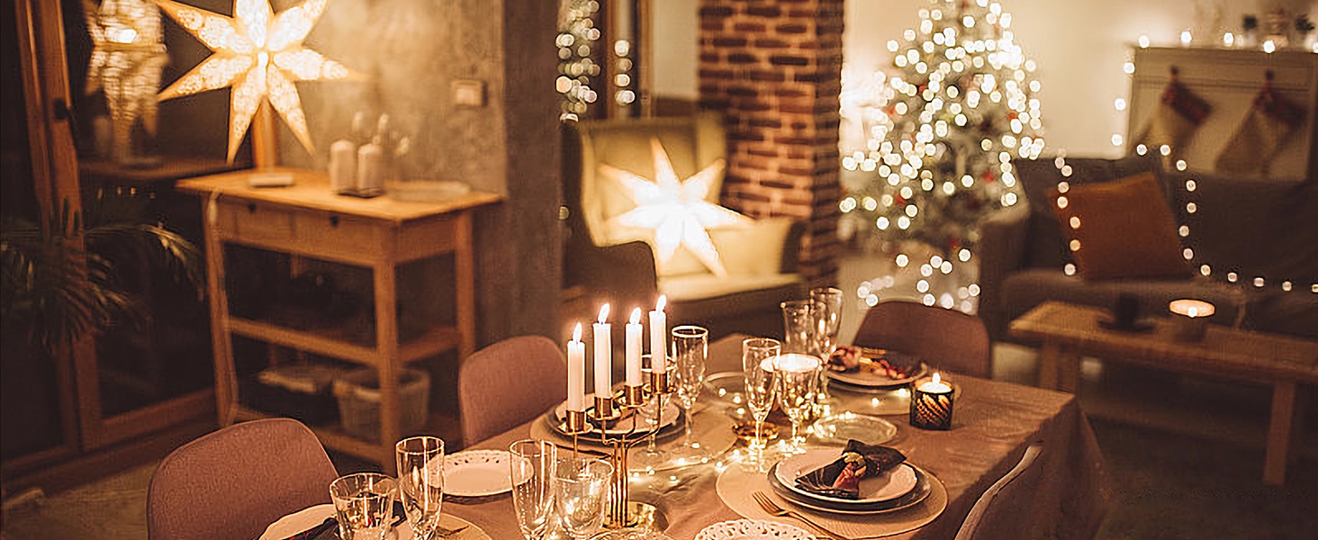 Festive dining table setting with lit candles, white star decorations, and string lights creating a warm holiday atmosphere. Place settings and glassware arranged elegantly.